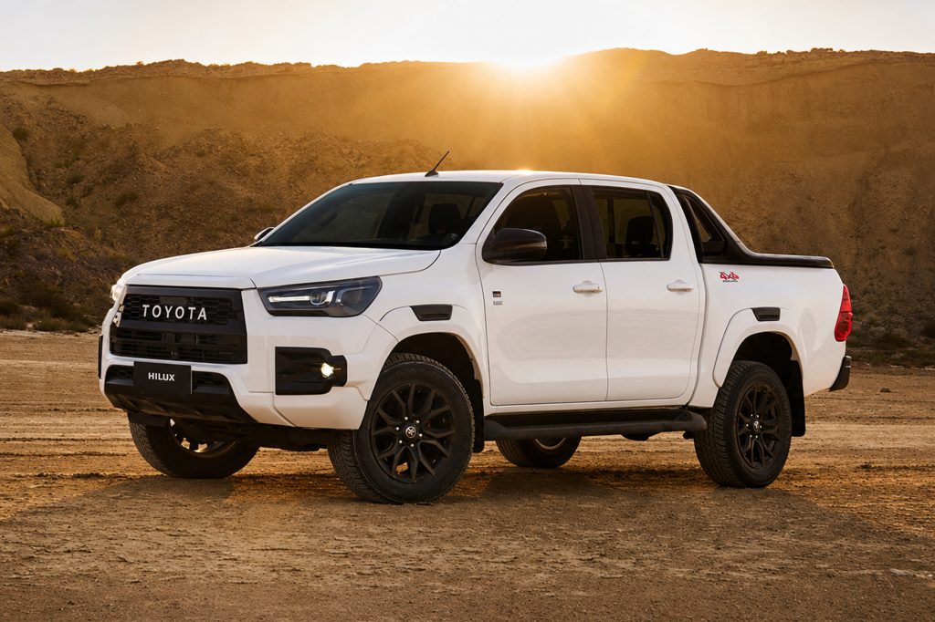 A white bulletproof Hilux parked on a dirt terrain at sunset, with rocky hills in the background.